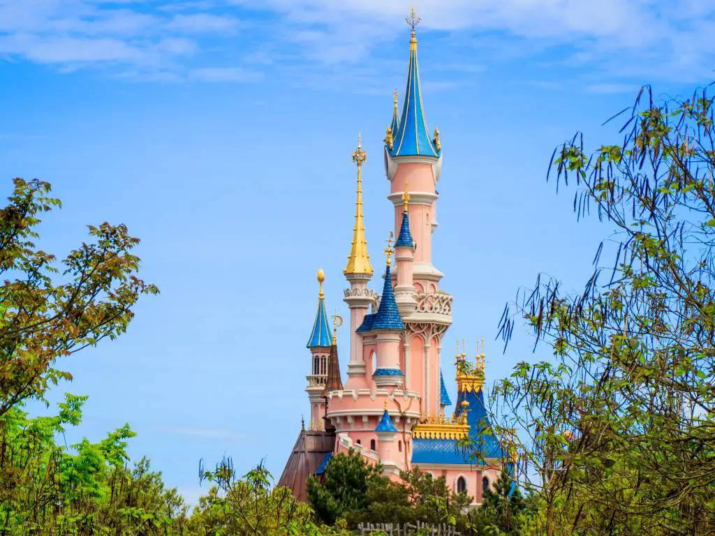 Pink and blue castle in Disneyland Paris, surrounded by green trees under the bright blue sky during daytime.