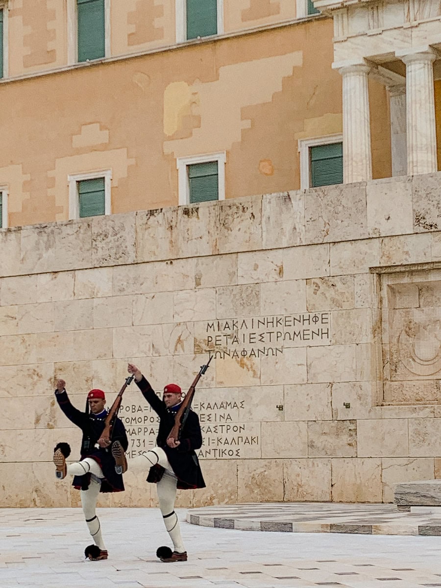 2 evzones raising their hand and leg in front of the monument of the unknown soldier.