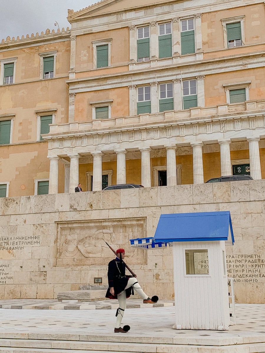 Presidential guard moving his leg up next to a white guard house with a blue roof.