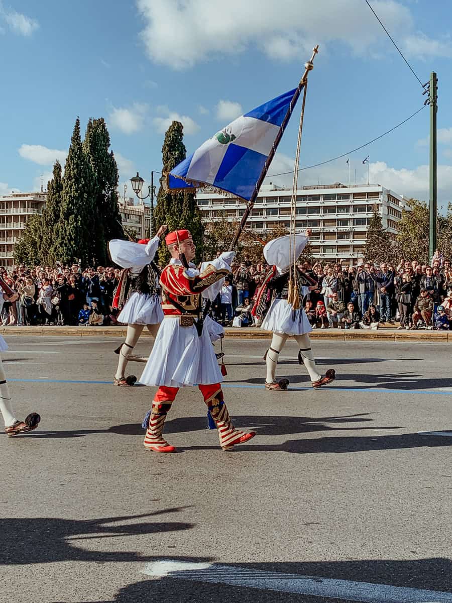Evzones marching by in their traditional outfits while holding the Greek flag.