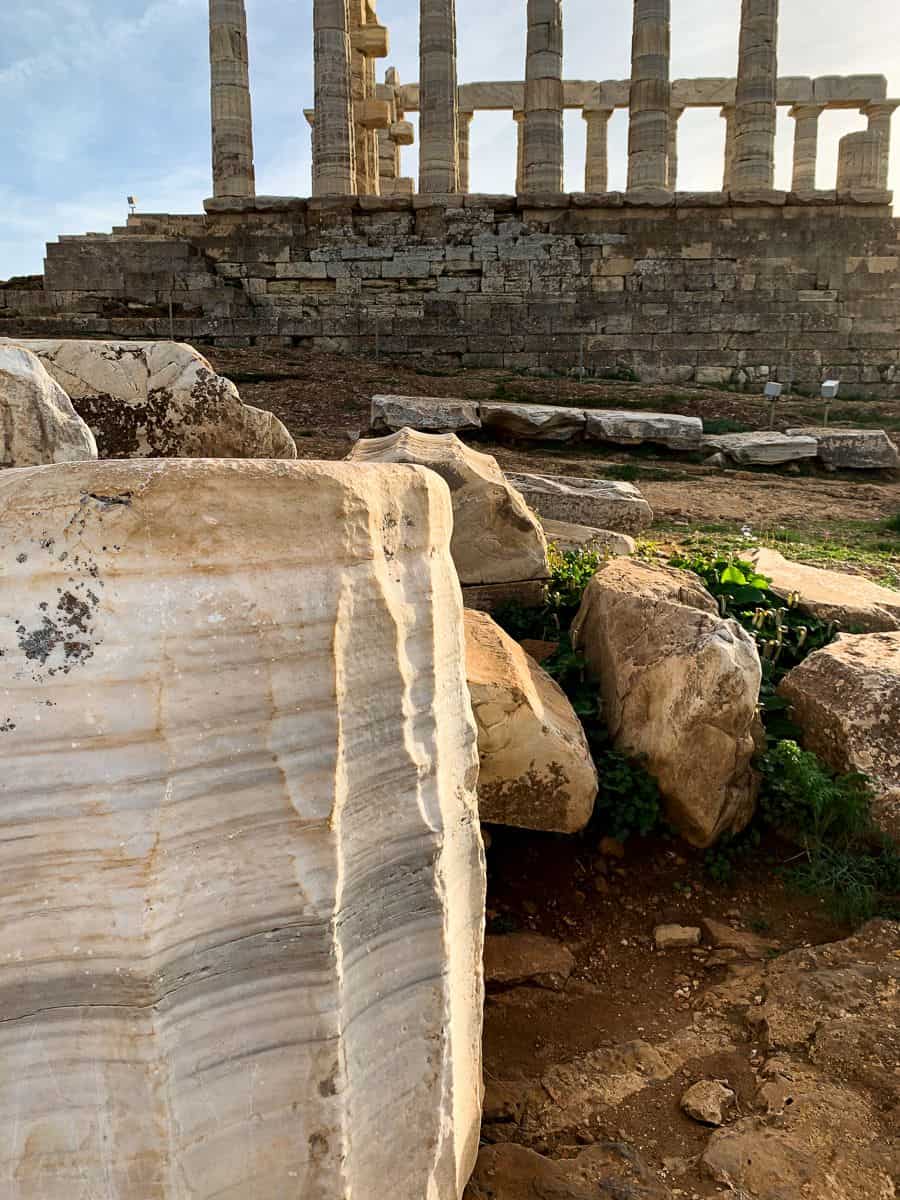 Large marble stone blocks lie in the foreground at the Temple of Poseidon at Cape Sounion with ancient columns rising behind them. The image highlights the scale, texture, and construction materials of the ruins, giving context to the temple’s original structure and age.