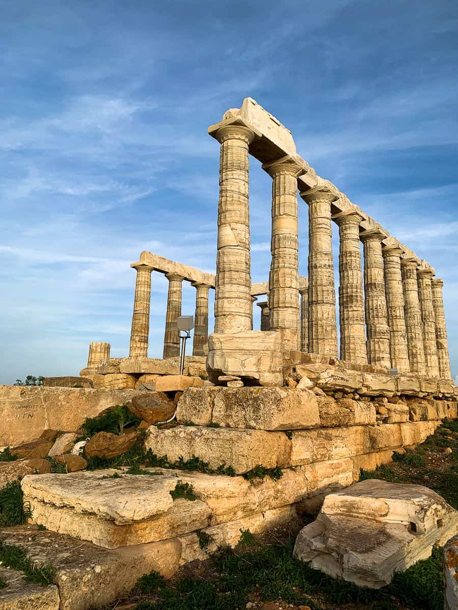 Ruins of the Temple of Poseidon with its tall white marble Doric columns standing on a rocky cliff overlooking the deep blue Aegean Sea under a clear sky.