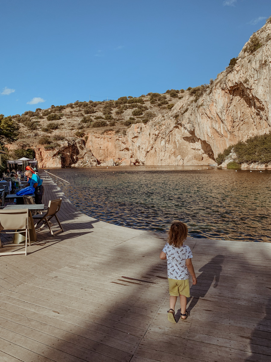 O standing by the dock at Lake Vouliagmeni surrounded by rocky mountains.