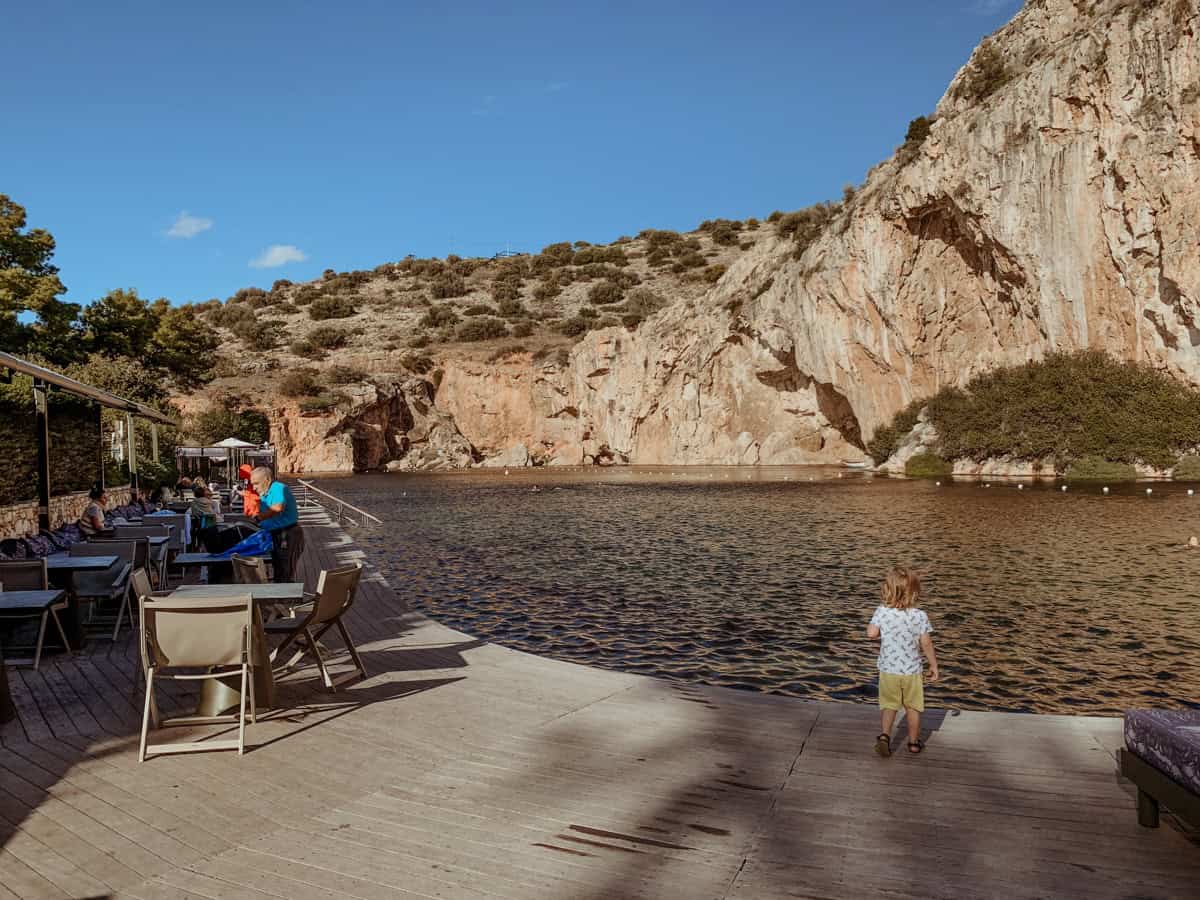 O standing by the dock at Lake Vouliagmeni, surrounded by rocky mountains.
