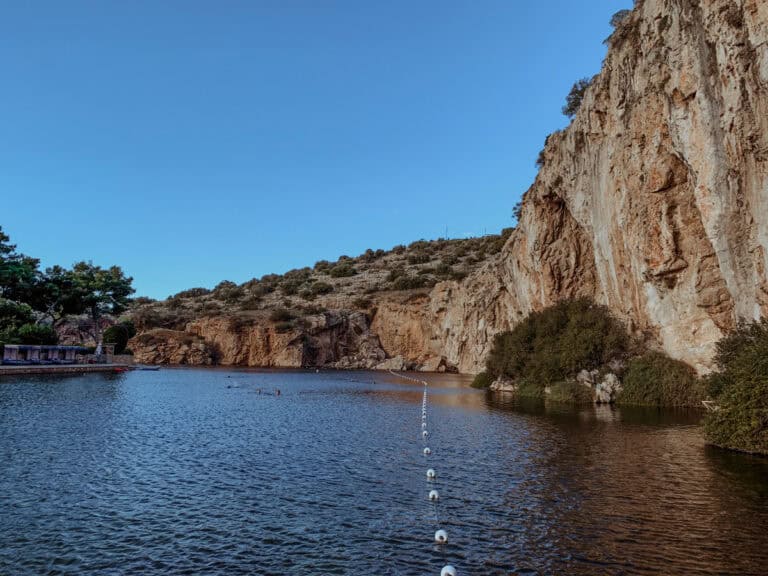 Lake Vouliagmeni with blue waters surrounded by rocky mountains under clear blue skies.