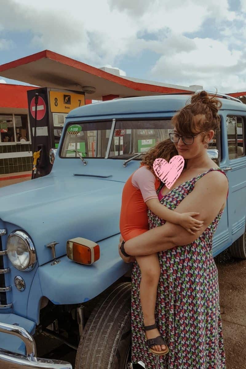 Babs is holding our daughter Norah at a Cuba gas station, with a Willys Jeep Station Wagon behind them.