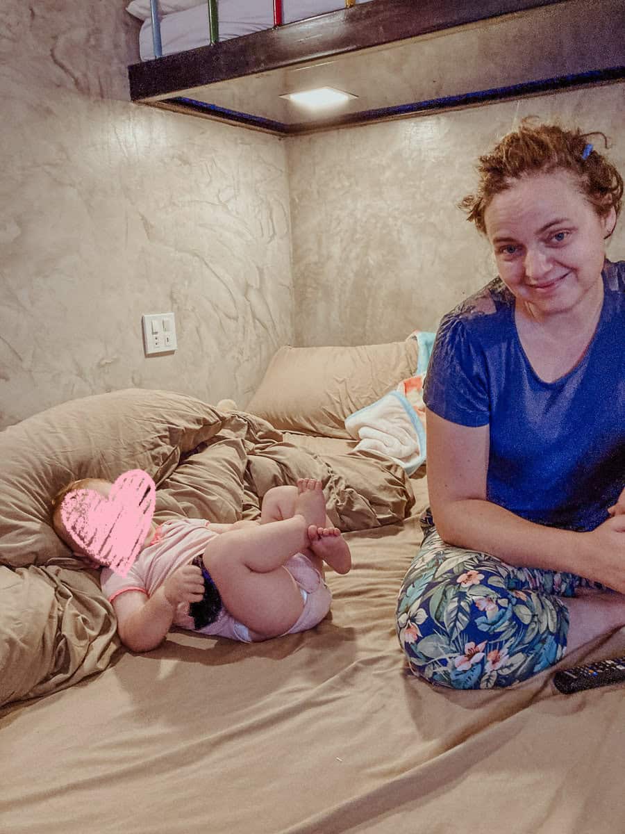 Kath sits cross-legged on the bed, smiling at the camera, while her baby relaxes on the bed.