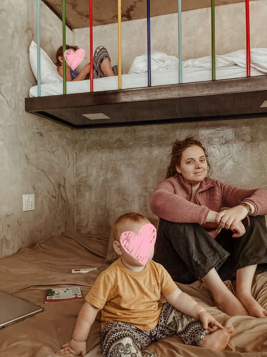 Kath sits on the bottom bunk in a mauve quarter-zip fleece and dark, wide-leg trousers, smiling gently at the camera. Oscar is sitting beside her wearing a mustard-yellow T-shirt, while Norah lies on the top bunk resting against a white pillow.