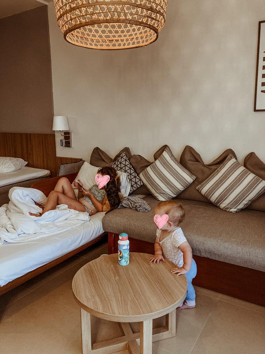 Norah lies on a white daybed, looking at her phone, while Oscar stands by a round wooden coffee table in the bright, warm living area of the hotel.