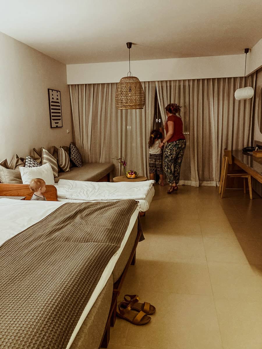 Kath stands by a window, helping Norah peek through tan curtains, while Oscar stands behind the bed looking toward them.