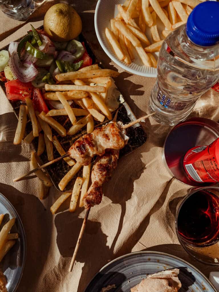 Greek street food at Isthmia Bridge Café with souvlaki skewers, fries, salad, pita, and drinks on a sunlit table.