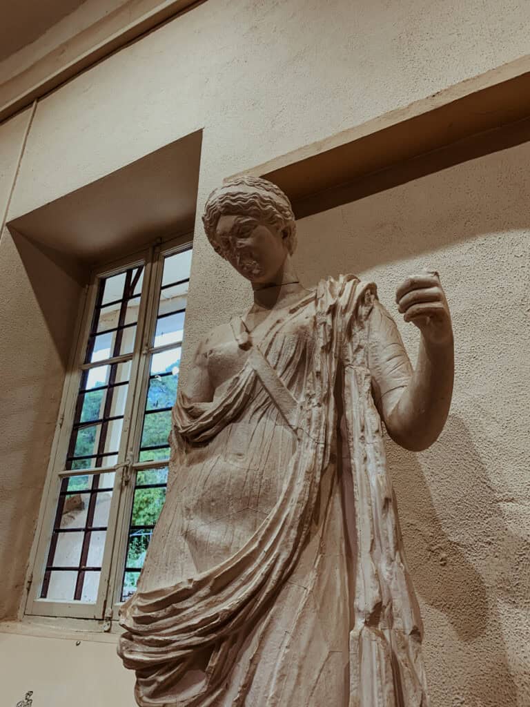 Close-up marble sculpture of a female figure (“Armed Aphrodite”) in a museum, wearing a draped chiton, with worn and cracked stone details, shown against a gallery wall with a window and trees.