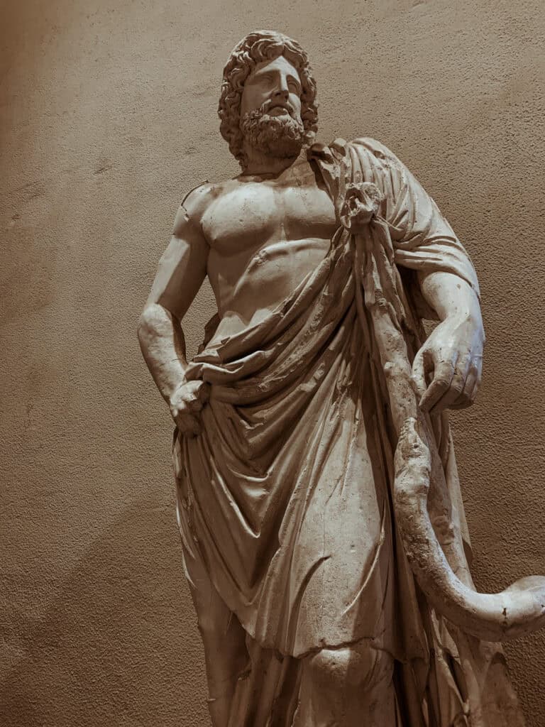 A weathered marble statue of Asclepius, the ancient Greek god of medicine, holding a thick, serpent-entwined staff.