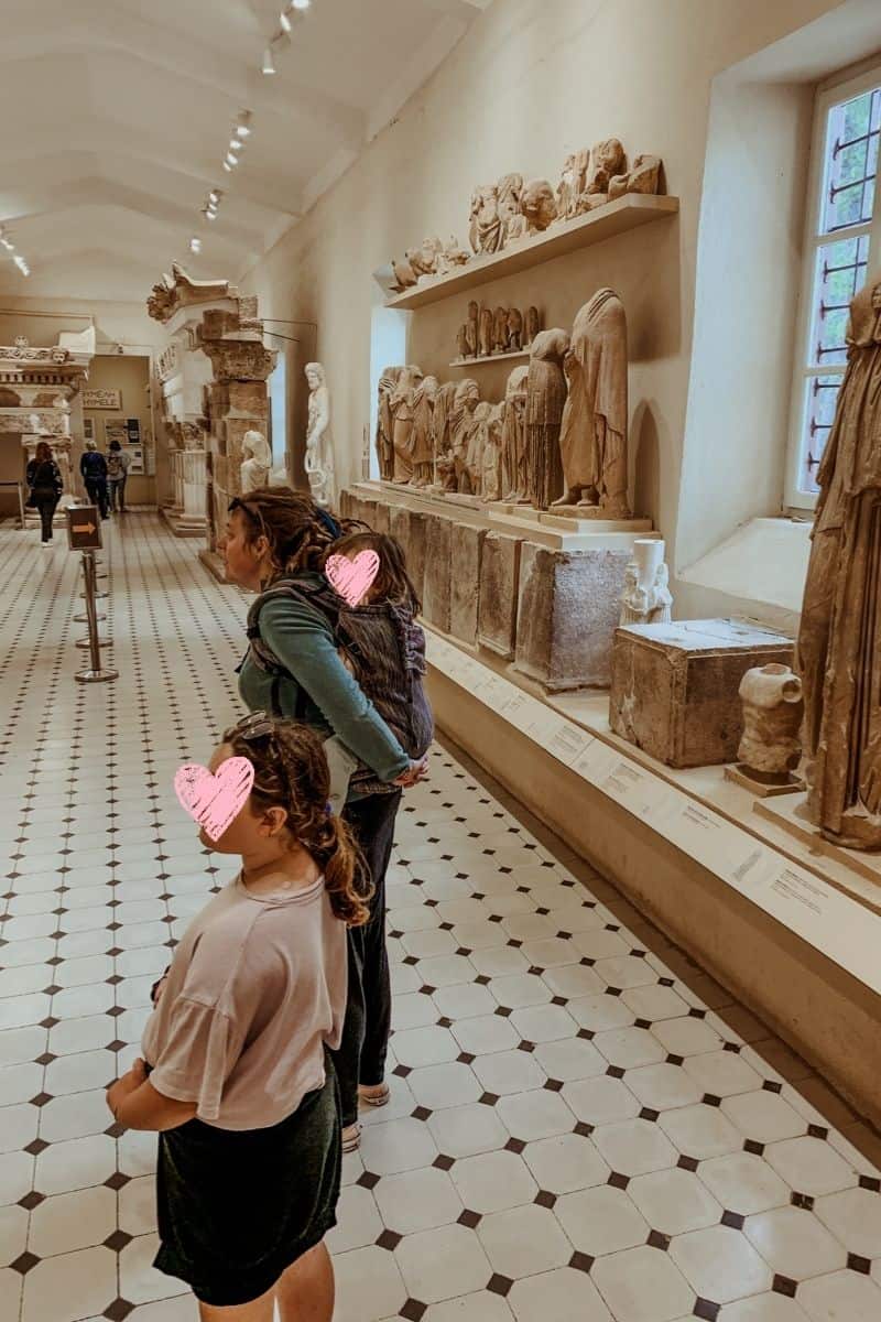 Kath piggybacked Oscar while Norah stood beside them, looking at the ancient stone statues and artifacts displayed in the Epidaurus Museum.