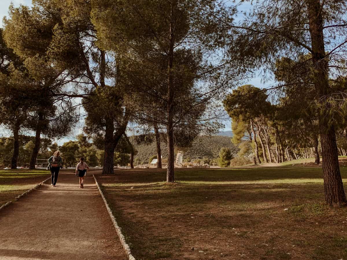 Kath with Oscar on her back and Norah beside them, walking along a dirt path lined with pine trees toward the archaeological site.