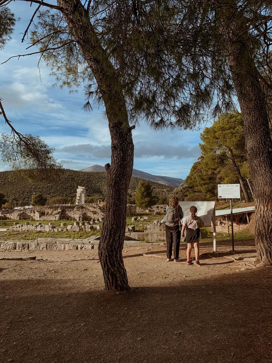 Kath with Oscar on her back and Norah beside them, standing under pine trees and looking at an information map in front of an ancient archaeological site with stone ruins and rolling hills.