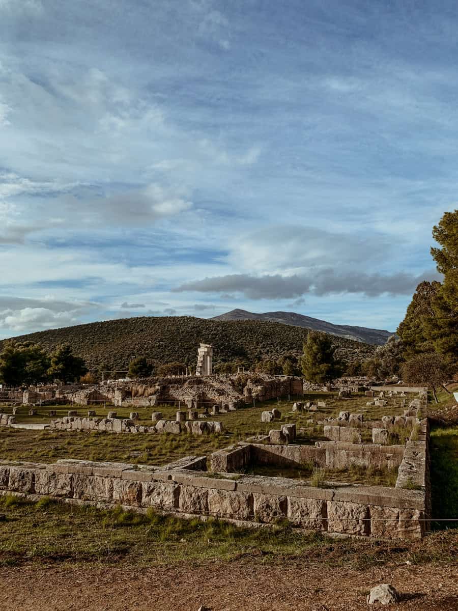 View of an ancient stone archaeological site with scattered ruins and a lone pillar structure, set against a backdrop of rolling hills and a cloudy sky.