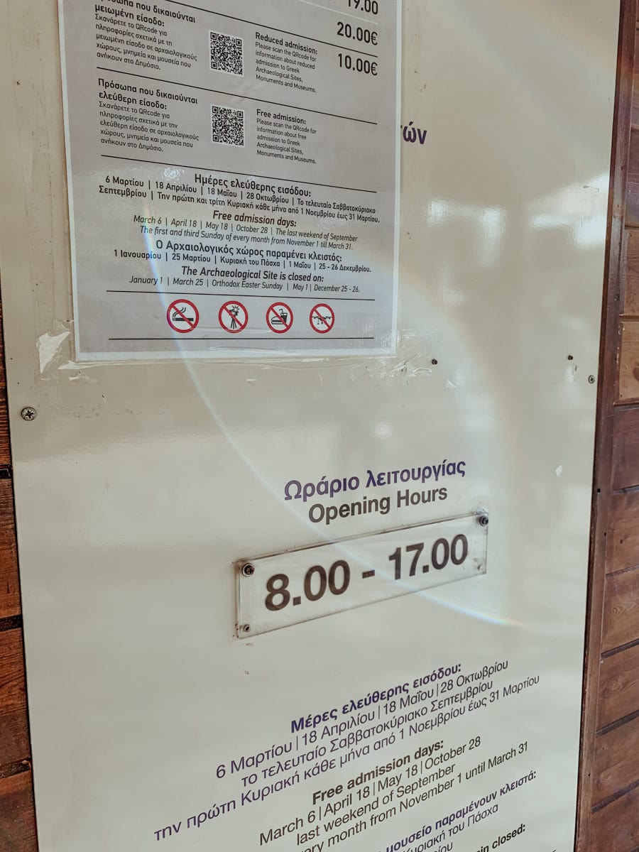 A close-up of printed signs displayed behind glass showing ticket prices, free admission days, closure dates, and opening hours (8.00 - 17.00) for an archaeological site.