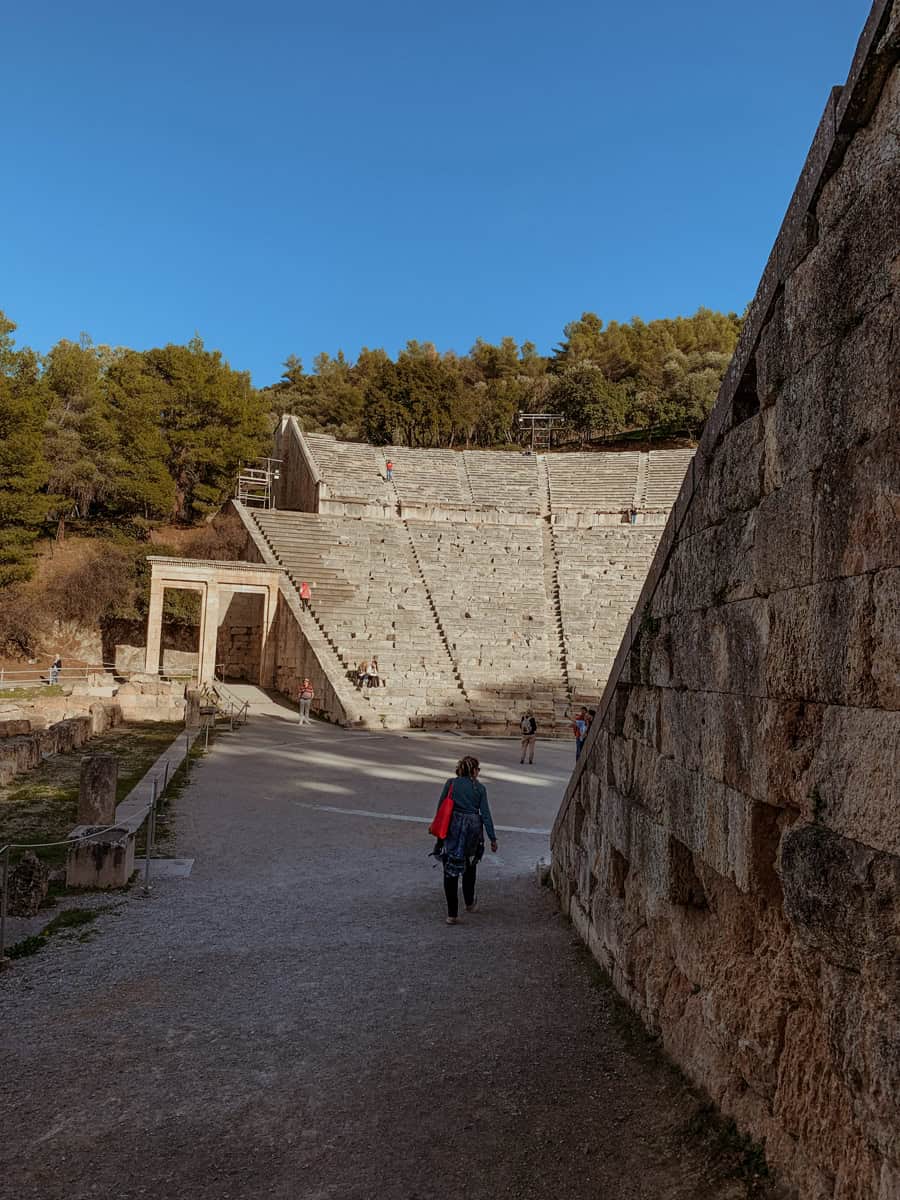Kath is walking along a gravel path toward the entrance and towering stone seating of the Ancient Theatre of Epidaurus under a clear blue sky.