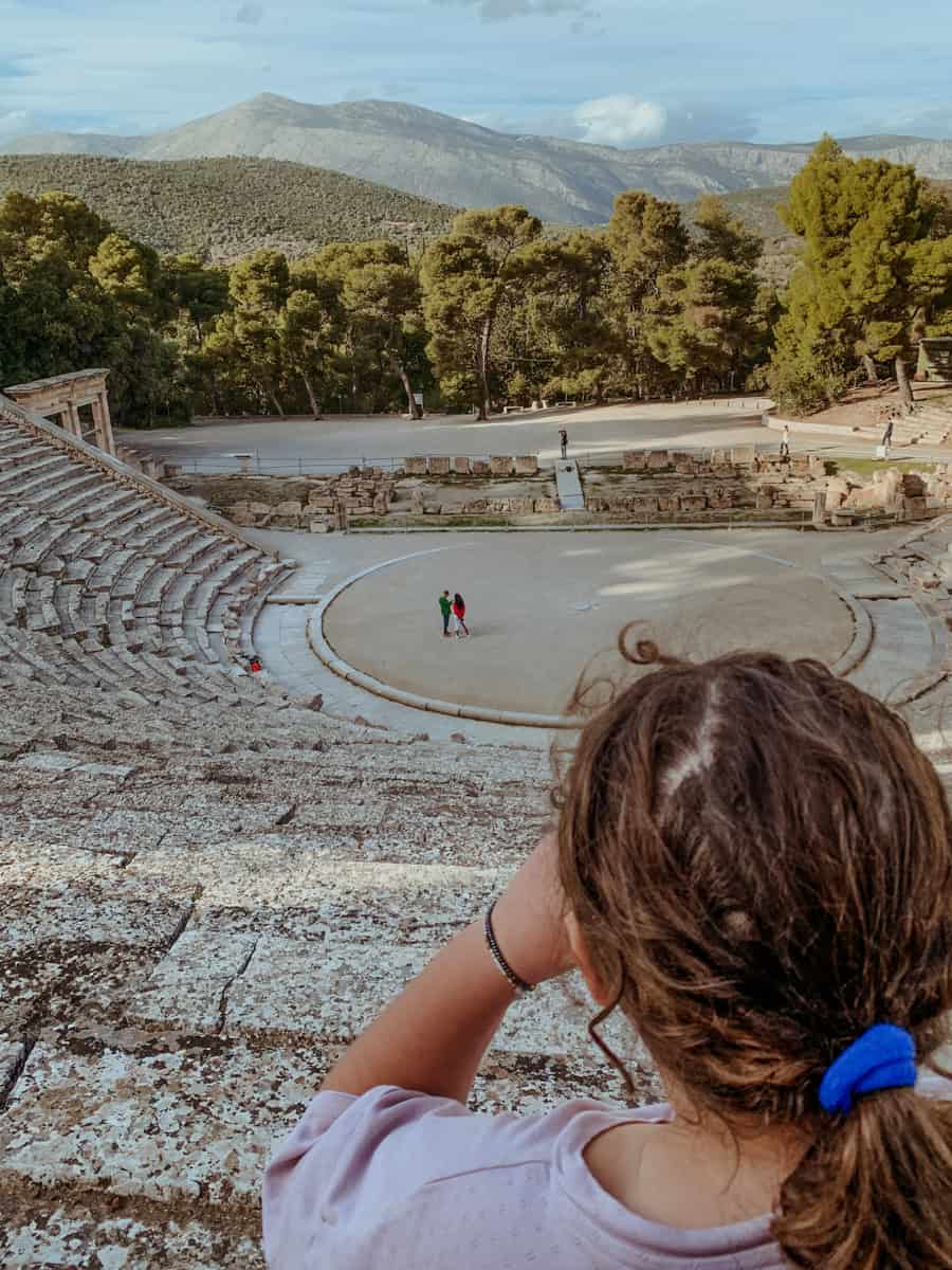 Norah looks down from the high stone seating onto the circular stage of a large ancient Greek amphitheater.
