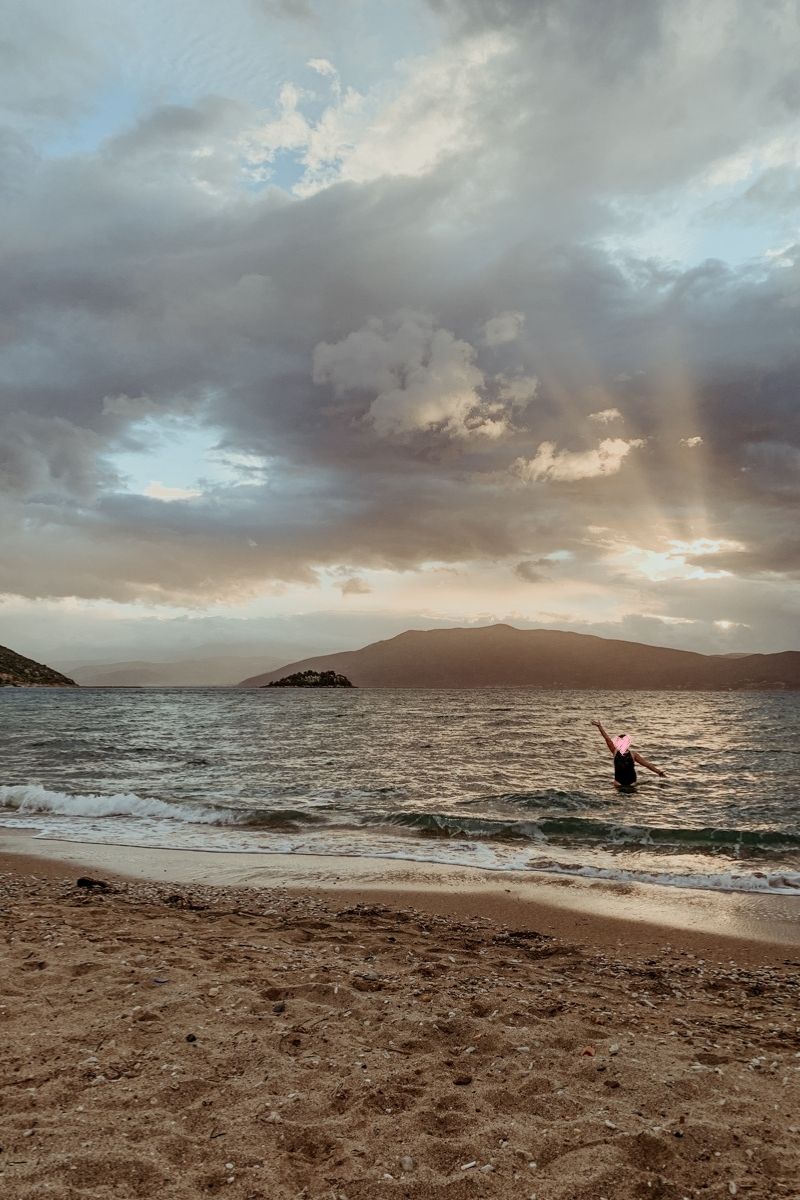 Norah stands waist-deep in the water at a sandy beach with one arm raised. Mountains and a cloudy sky are in the background.