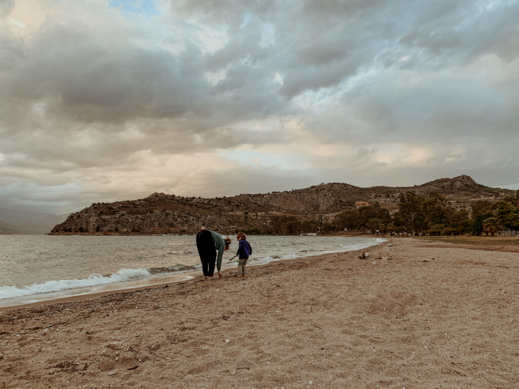 Kath bends down by the shoreline while Oscar stands next to her holding a stick on a wide, sandy beach.