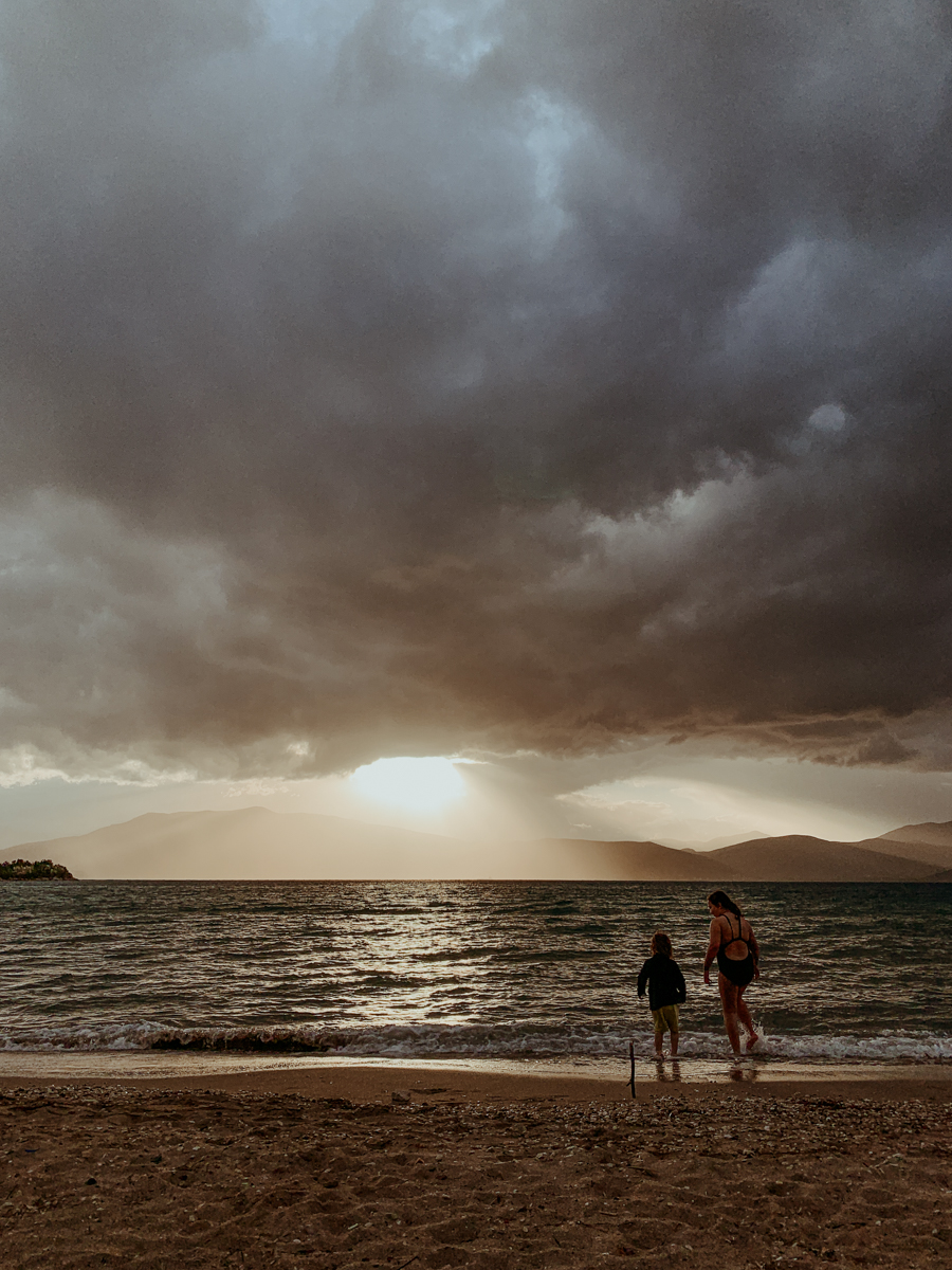 Norah and Oscar stand by the water on a sandy beach at sunset.