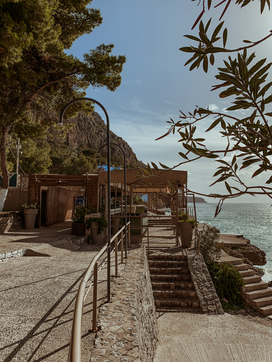 A paved walkway and stone stairs lead toward a coastal restaurant overlooking the ocean at Paralia Arvanitias.