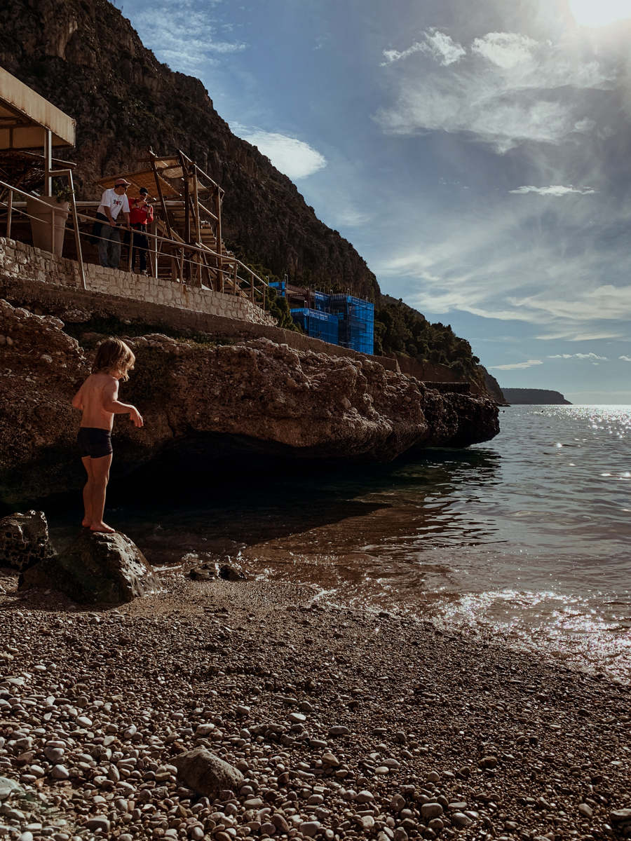 Oscar stands on a rock at the shoreline of Paralia Arvanitias near a rocky cliffside structure.