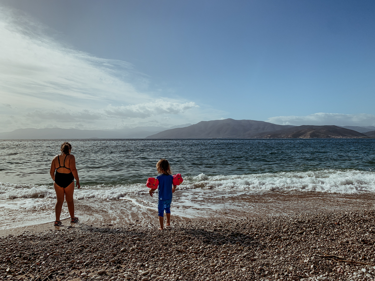 Norah, wearing a black swimsuit, and Oscar, wearing a blue swim shirt and orange arm floaties, stand on the pebble shore of Paralia Arvanitias.