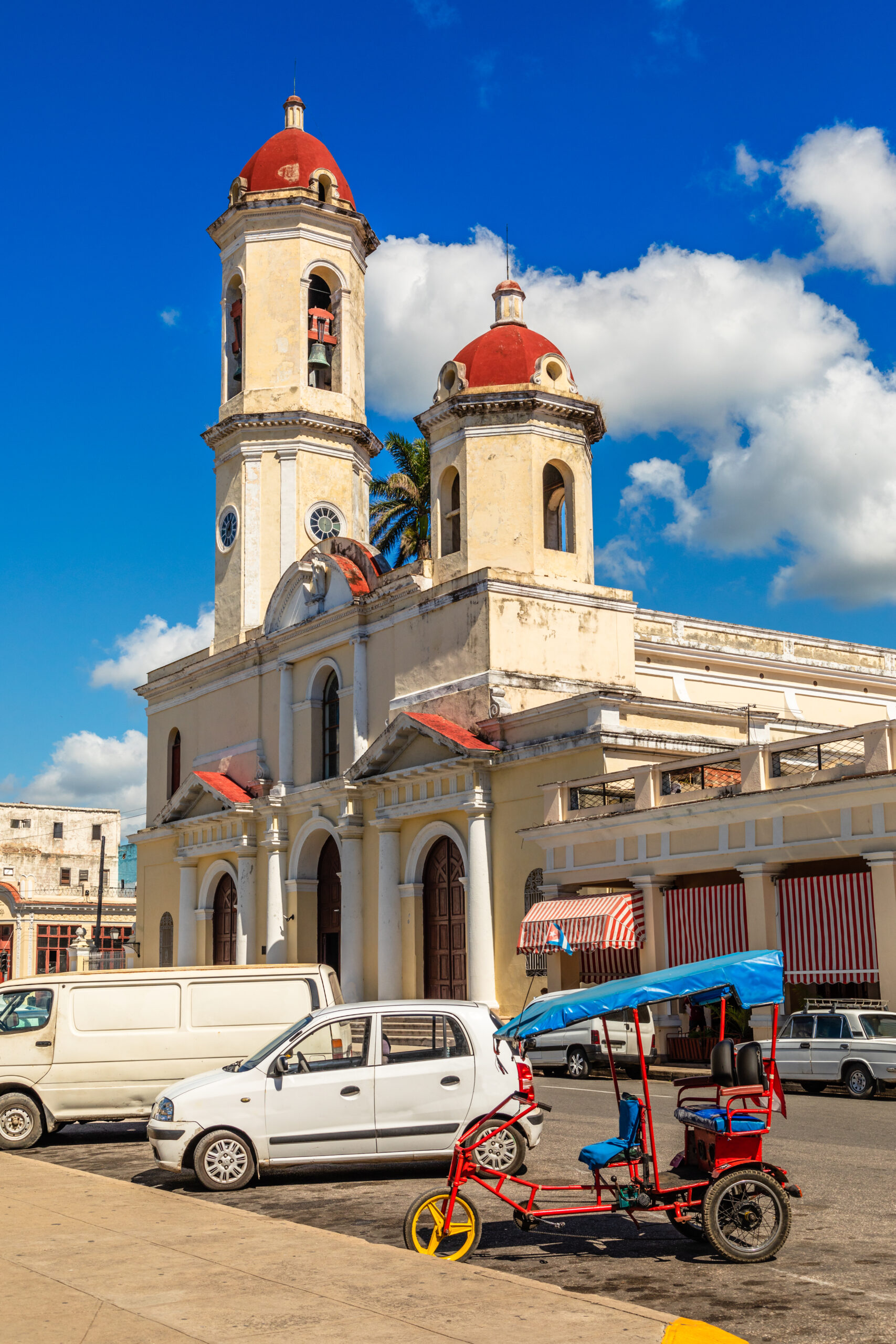 A view of Cienfuegos Cathedral in Cienfuegos, Cuba, with a red bicitaxi and a white car parked in front.