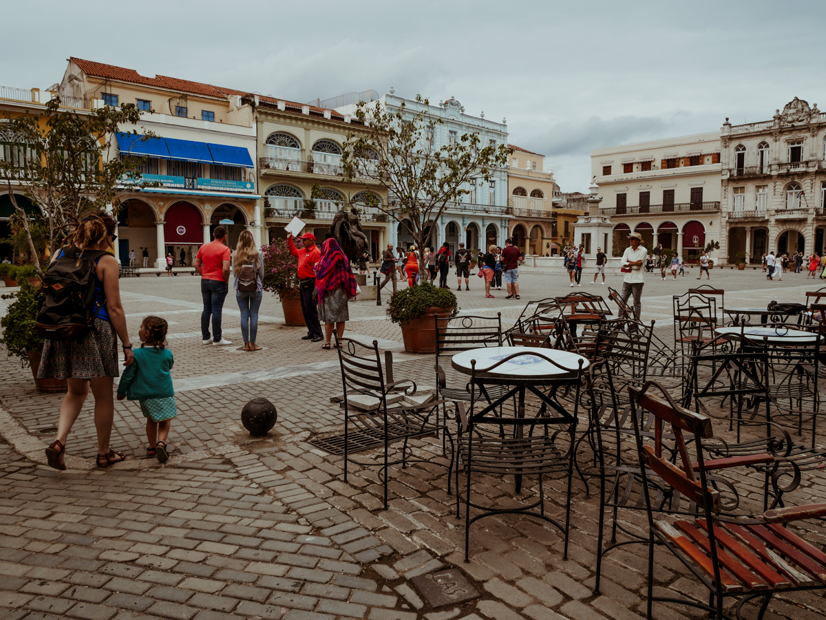 Kath and Norah walk past empty iron tables and chairs at an outdoor restaurant in Plaza Vieja, a historic cobblestone square in Old Havana, surrounded by colonial buildings and a few tourists.