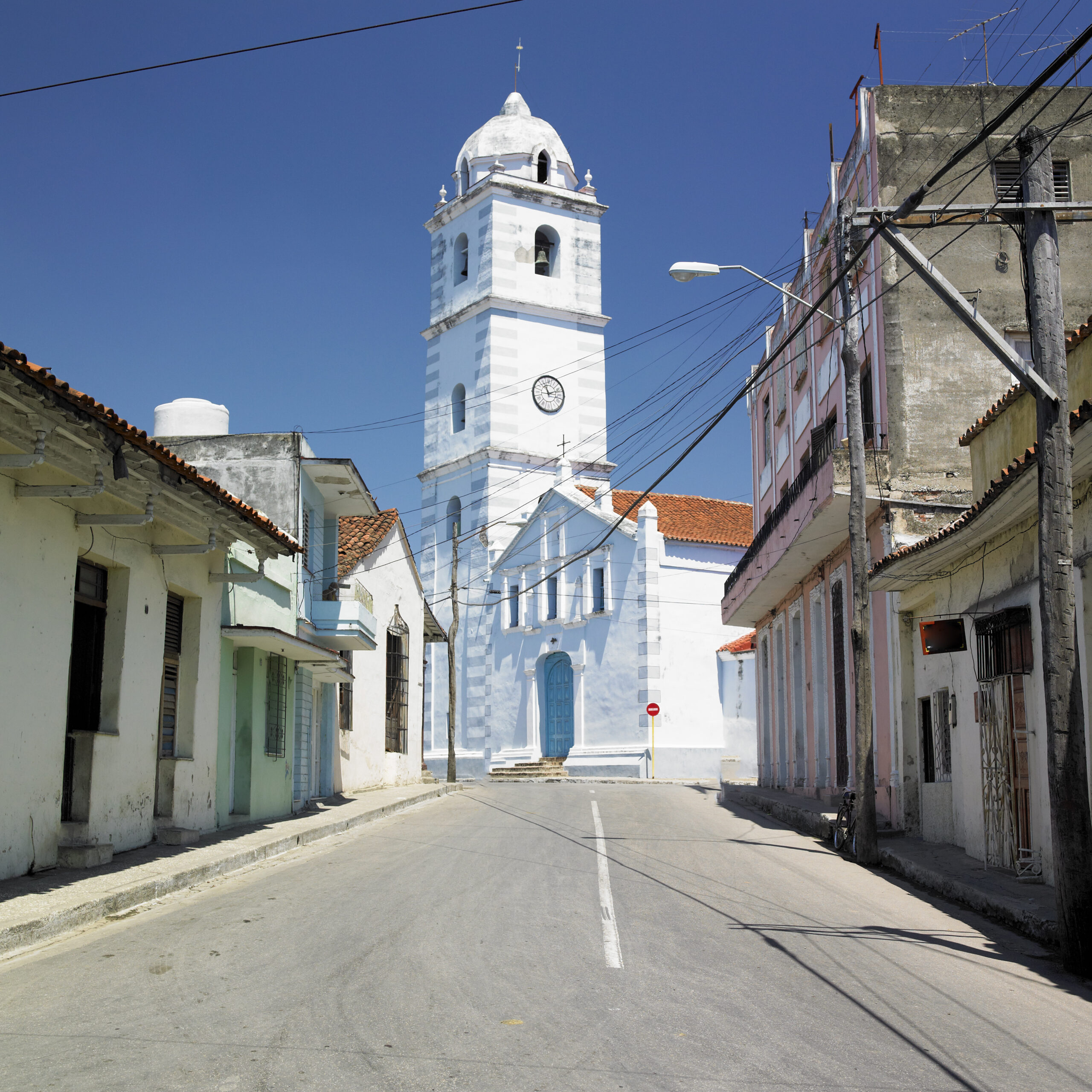 A paved street leading to Iglesia Parroquial Mayor del Espíritu Santo in Sancti Spíritus, Cuba, lined with colorful low-rise buildings.