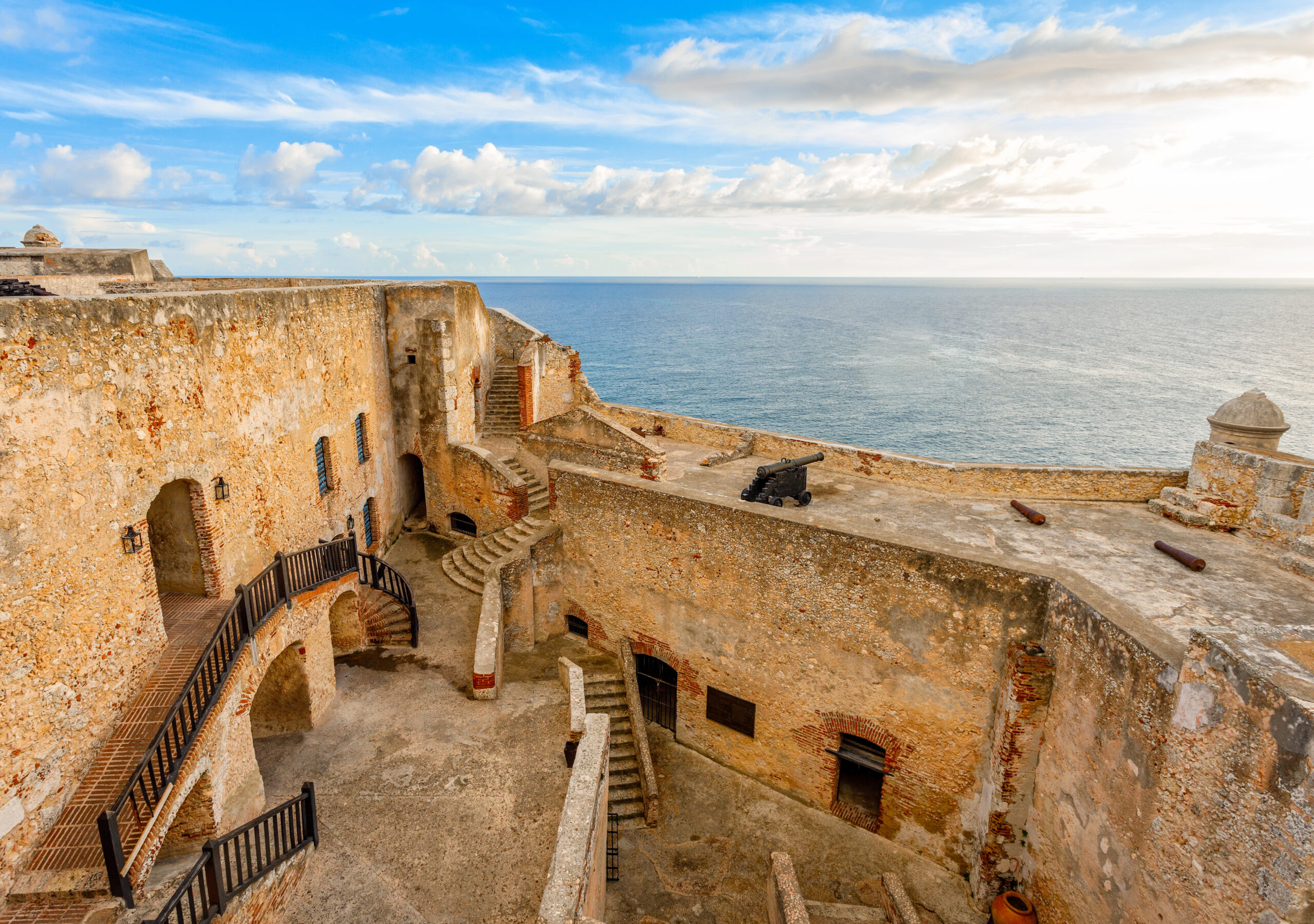 An interior view of Castillo de San Pedro de la Roca in Santiago de Cuba, with historic cannons, a weathered stone courtyard, and multi-level brick staircases.